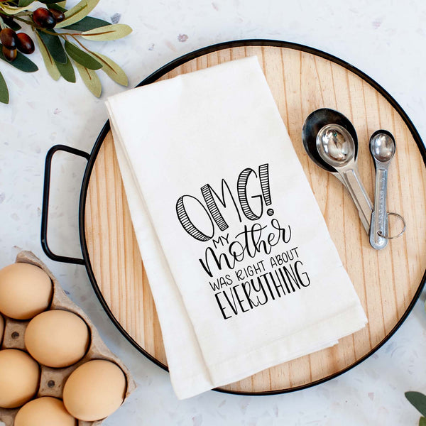 White floursack kitchen towel with black hand lettered illustrated design that says OMG! My mother was right about everything shown folded on a serving tray with a set of measuring spoons and fresh eggs