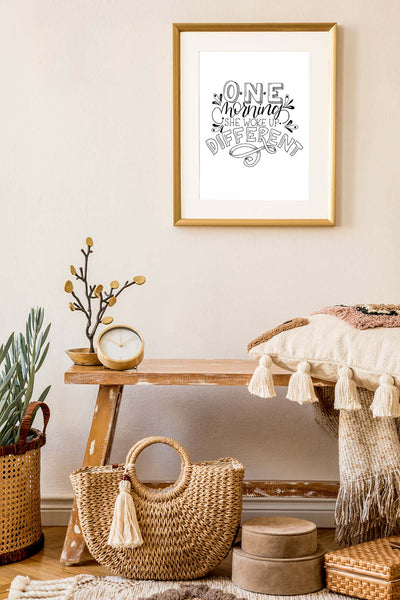 Original black and white illustrated wall art that says one morning she woke up different shown hanging on a wall in a living room with a bench pillow and accessories