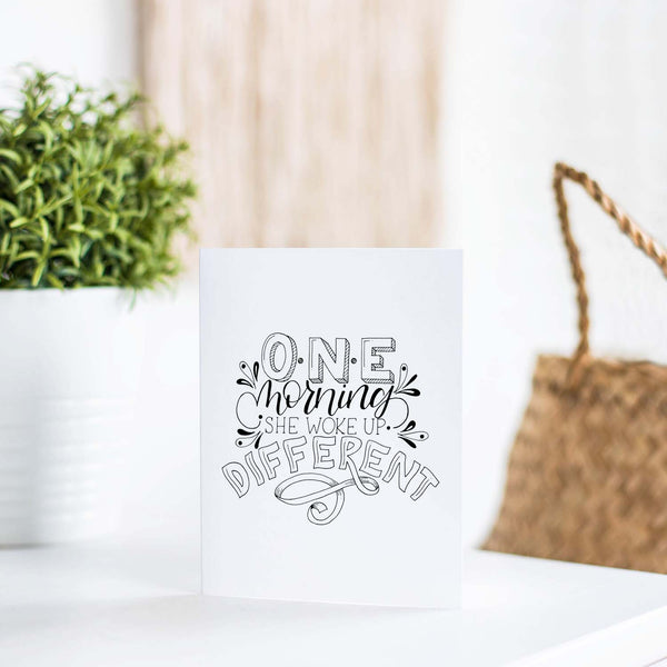 one morning she woke up different hand lettered and illustrated black and white greeting card with A2 envelope on a white table with a plant and handbag