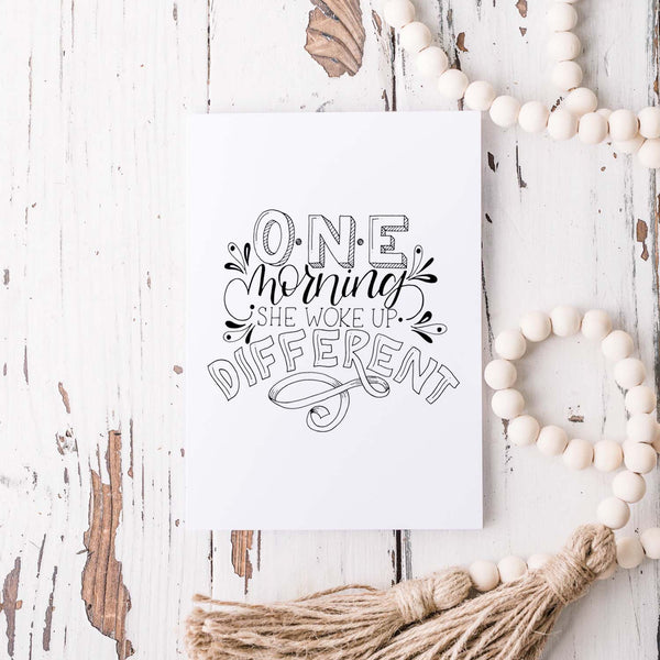 one morning she woke up different hand lettered and illustrated black and white greeting card with A2 envelope shown on a white rustic table with a white wooden bead garland