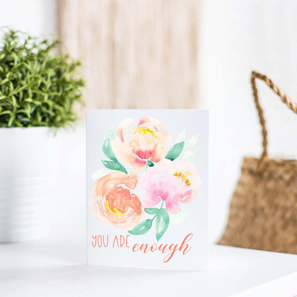 watercolor peonies in soft pastel colors friendship greeting card that says you are enough with a white A2 envelope shown standing on a white table with a plant and handband