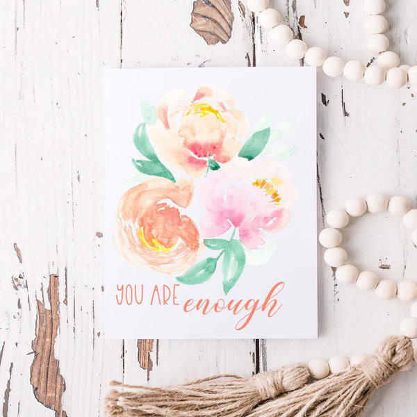 watercolor peonies in soft pastel colors friendship greeting card that says you are enough with a white A2 envelope shown laying on a rustic white wood table with a white wooden bead gardland