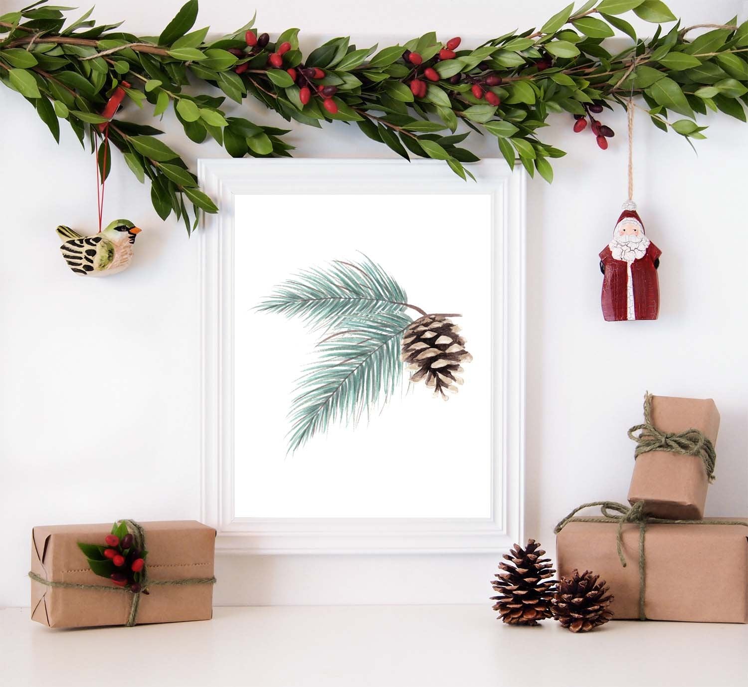 Watercolor painting of evergreen branches and pinecone shown in a white frame surrounded by christmas decorations