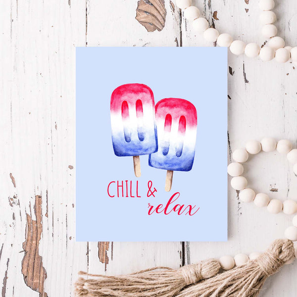watercolor red white and blue summertime popsicles on a friendship greeting card that says chill & relax with a white A2 envelope shown laying on a rustic white wooden table with a white wood bead garland