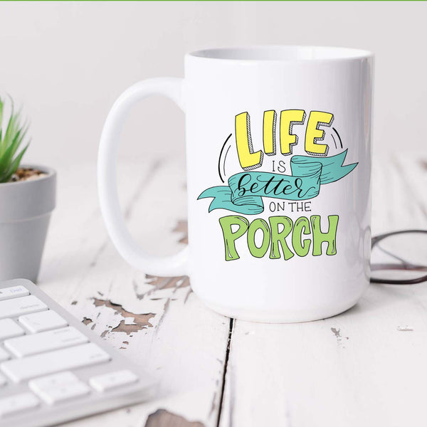 15oz white ceramic mug with hand lettered illustrated design that says Life Is Better On The Porch shown on a desk with plant, keyboard and glasses