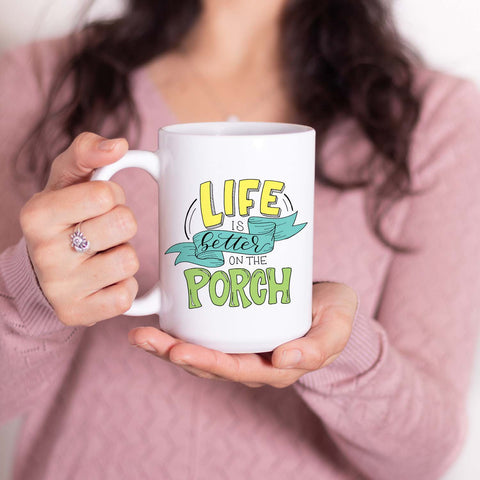15oz white ceramic mug with hand lettered illustrated design that says Life Is Better On The Porch shown held by a woman