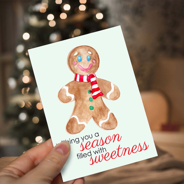 Watercolor Christmas card with a happy watercolor gingerbread man that says wishing you a season filled with sweetness shown with a woman holding in front of a lighted Christmas tree