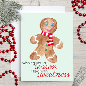 Watercolor Christmas card with a happy watercolor gingerbread man that says wishing you a season filled with sweetness shown with holiday decorations