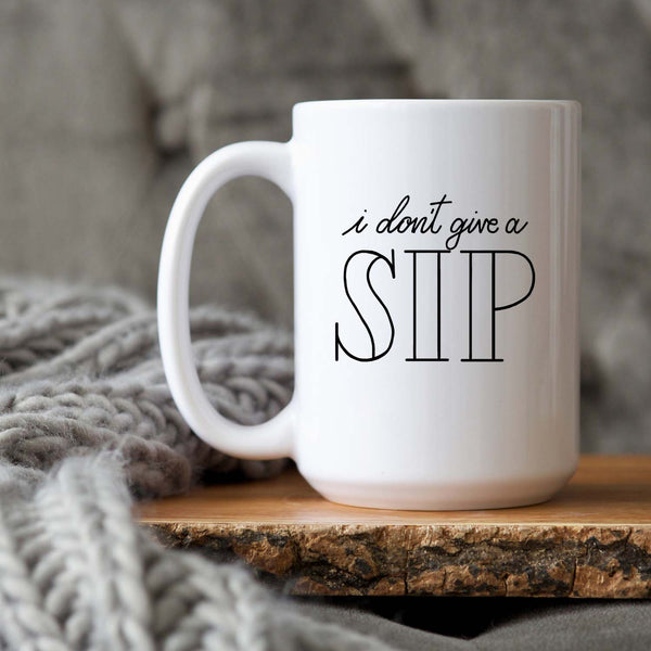 15oz white ceramic mug with hand lettered illustrated design that says I don't give a sip shown sitting on a wood tray with a grey knit blanket