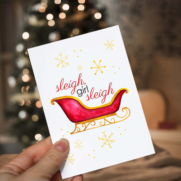 Watercolor Christmas greeting card with a watercolor painted red and gold sleigh and gold snowflakes with message that says sleigh, girl sleigh shown with a woman holding card in front of lighted christmas tree