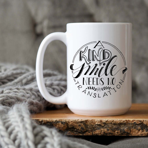 15oz white ceramic mug with hand lettered illustrated design that says a kind smile needs no translation shown on a wood tray with a grey knit blanket