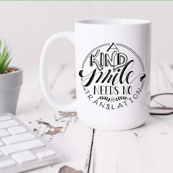 15oz white ceramic mug with hand lettered illustrated design that says a kind smile needs no translation shown on a desk with a plant, keyboard, glasses