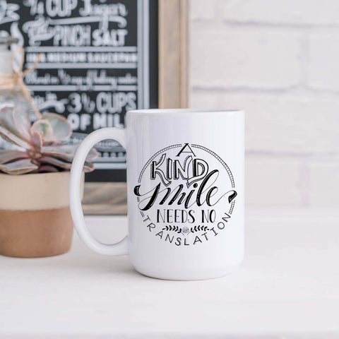 15oz white ceramic mug with hand lettered illustrated design that says a kind smile needs no translation shown in a kitchen