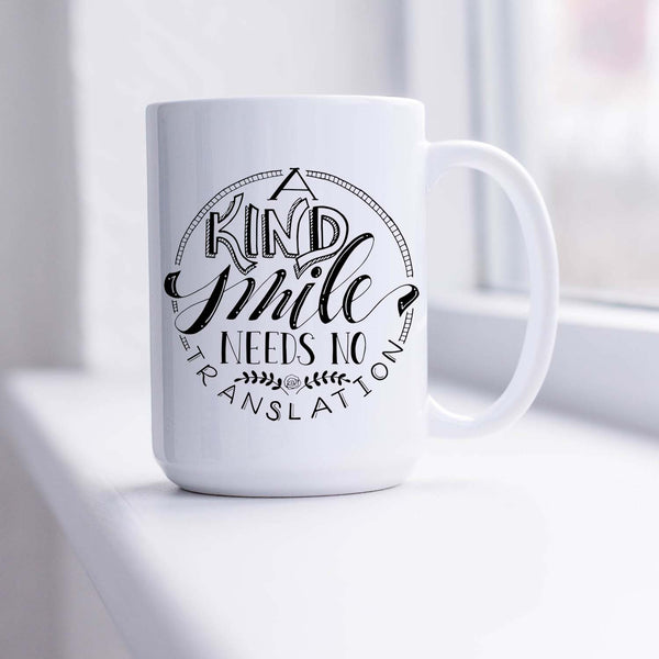 15oz white ceramic mug with hand lettered illustrated design that says a kind smile needs no translation sitting in a sunny window