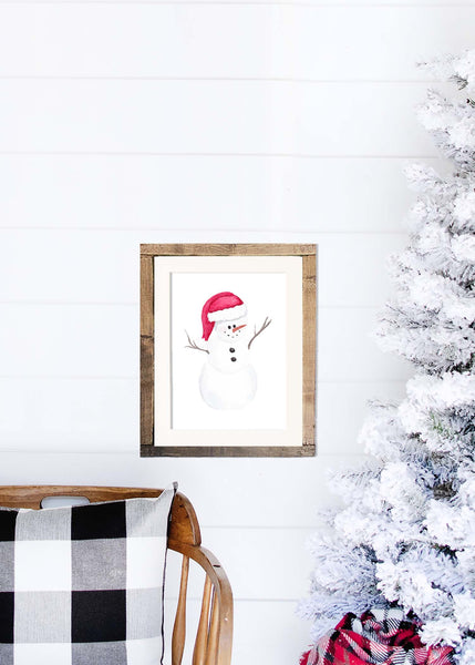 Watercolor painting of a chubby snowman wearing a santa hat shown in wood frame hanging on a shiplap wall next to a white flocked tree and rocking chair