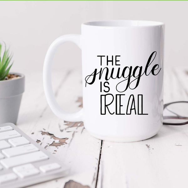 15oz white ceramic mug with hand lettered illustrated design that says the snuggle is real shown sitting on white office desk