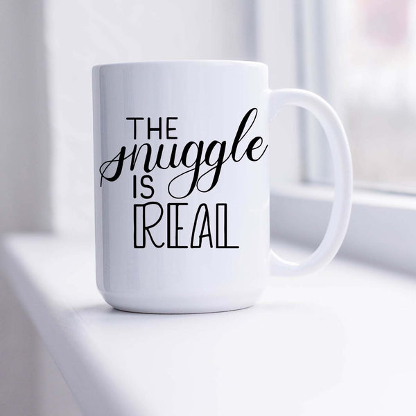 15oz white ceramic mug with hand lettered illustrated design that says the snuggle is real shown sitting in a sunny window