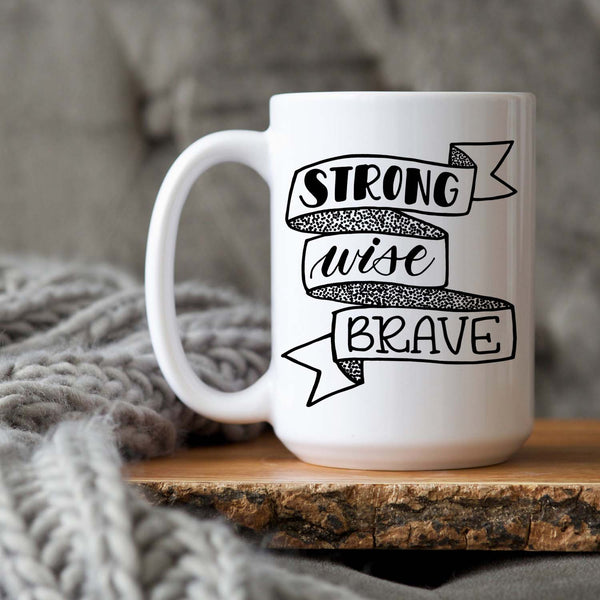 15oz white ceramic mug with hand lettered illustrated design that says strong wise brave inside a ribbon illustration shown sitting on a wood tray with a grey knit blanket