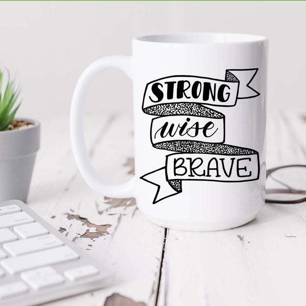 15oz white ceramic mug with hand lettered illustrated design that says strong wise brave inside a ribbon illustration sitting on a white office desk