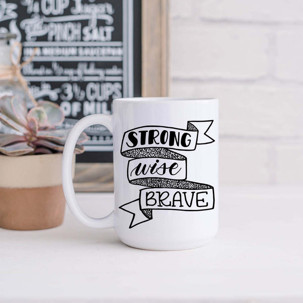 15oz white ceramic mug with hand lettered illustrated design that says strong wise brave inside a ribbon illustration shown in a kitchen