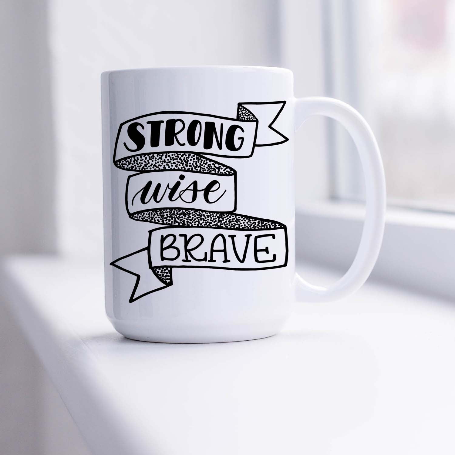 15oz white ceramic mug with hand lettered illustrated design that says strong wise brave inside a ribbon illustration sitting in a sunny window