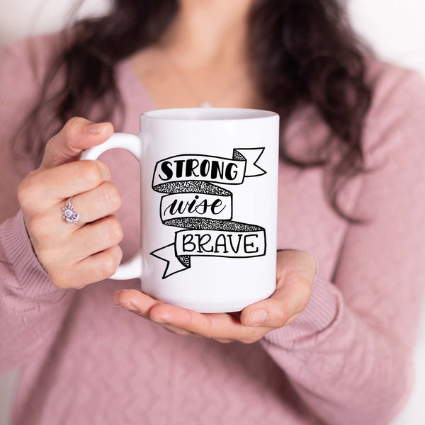 15oz white ceramic mug with hand lettered illustrated design that says strong wise brave inside a ribbon illustration shown with a woman holding the mug