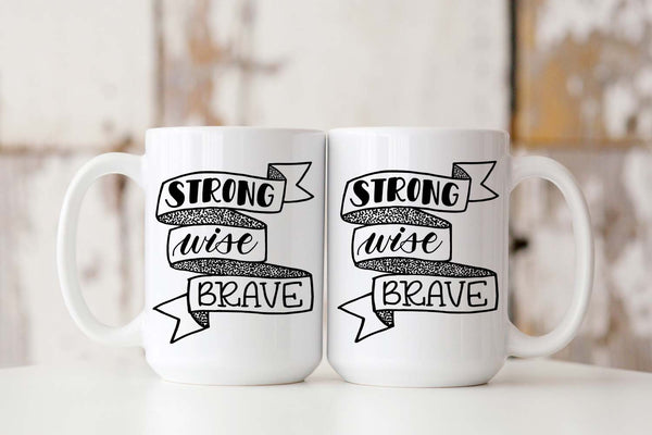 15oz white ceramic mug with hand lettered illustrated design that says strong wise brave inside a ribbon illustration showing front and back of mug