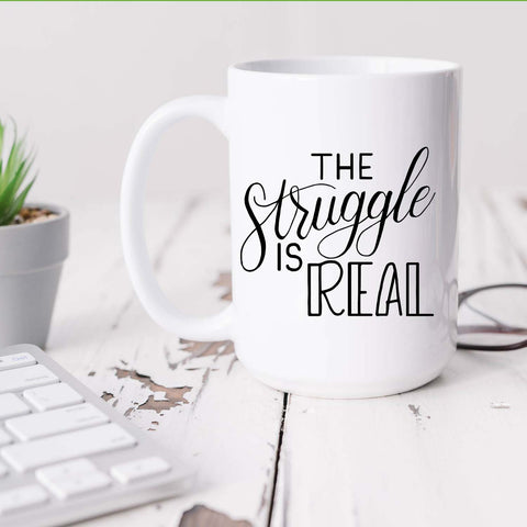 15oz white ceramic mug with hand lettered illustrated design that says the struggle is real shown sitting on a white office desk