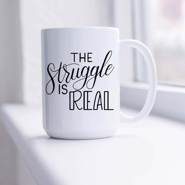 15oz white ceramic mug with hand lettered illustrated design that says the struggle is real shown sitting in a sunny window