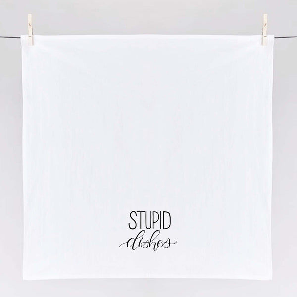 White floursack towel with black hand lettered illustrated design that says Stupid dishes shown unfolded and hanging from clothes pins