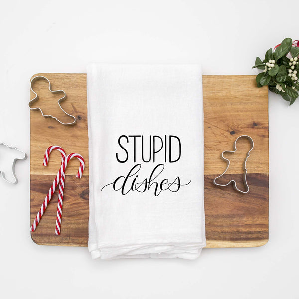 White floursack towel with black hand lettered illustrated design that says Stupid dishes shown folded on a wood cutting board with Christmas cookie cutters and candy canes