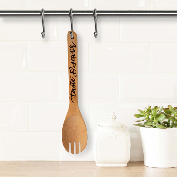 Bamboo round serving fork that says taste & savor in burned hand lettering shown hanging in a kitchen next to a canister and potted plant