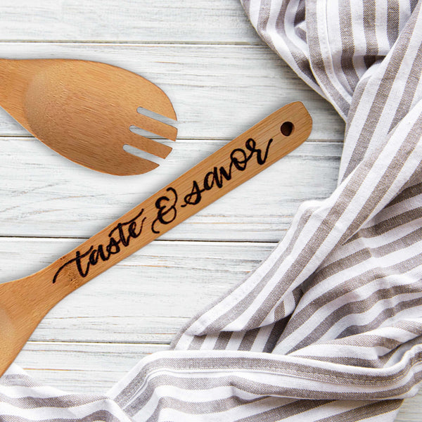 Bamboo round serving fork that says taste & savor in burned hand lettering laying on a table with a towel