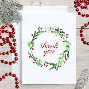 Watercolor Christmas greeting card with a holiday wreath of twigs, greens and berries in red and green that says thank you shown with holiday decorations