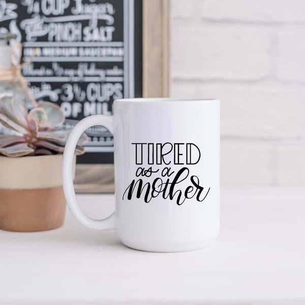 15oz white ceramic mug with hand lettered illustrated design that says tired as a mother shown sitting in a kitchen