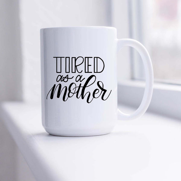 15oz white ceramic mug with hand lettered illustrated design that says tired as a mother shown sitting in a sunny window