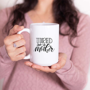 15oz white ceramic mug with hand lettered illustrated design that says tired as a mother shown with a woman holding the mug