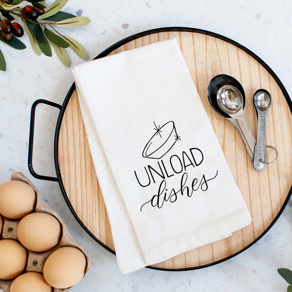 White floursack towel with black hand lettered illustrated design that says Unload dishes with a clean bowl doodle shown folded on a serving tray with a set of measuring spoons and fresh eggs