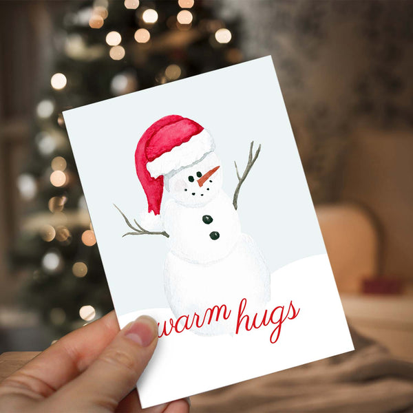 Watercolor Christmas greeting card with cute painted snowman with a red santa hat that says warm hugs shown with a woman holding card in front a lighted christmas tree