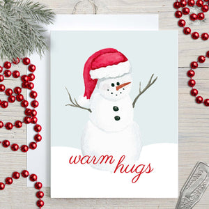 Watercolor Christmas greeting card with cute painted snowman with a red santa hat that says warm hugs shown with holiday decorations