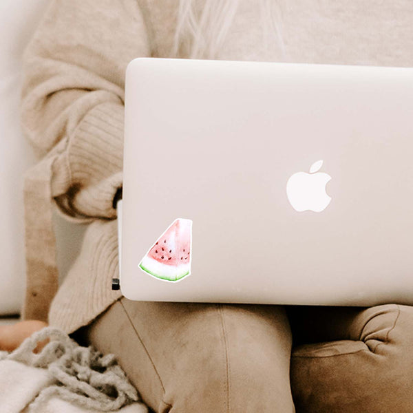 3" vinyl sticker of a watercolor painted slice of watermelon shown adhered to a MacBook cover sitting open on a woman's lap