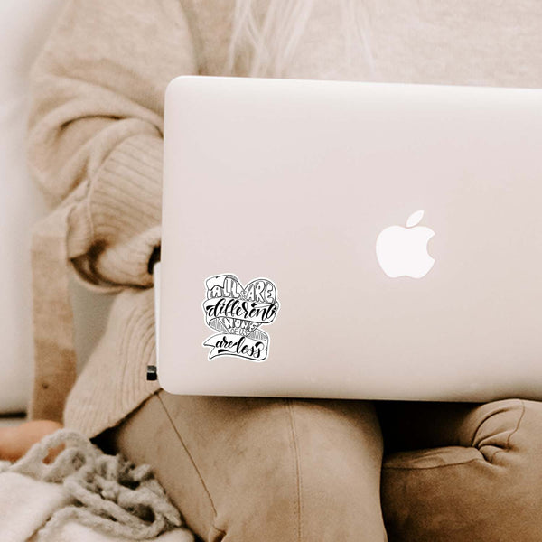 3" hand lettered, illustrated, black and white vinyl sticker saying all of us are different none of us are less shown adhered on a MacBook laptop cover sitting open on a woman's lap
