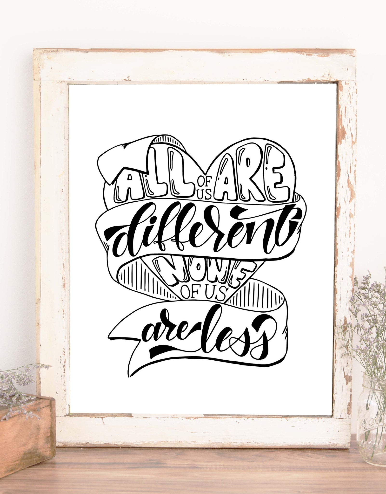 hand lettered wall art design in black and white that says all of us are different none of us are less with an illustration of a heart and winding ribbon shown in a rustic frame on a shelf with accessories