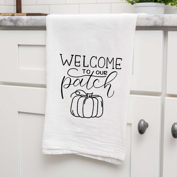 White floursack towel with black hand lettered illustrated design that says Welcome to our patch with pumpkin doodle shown folded and hanging from a countertop in a modern kitchen