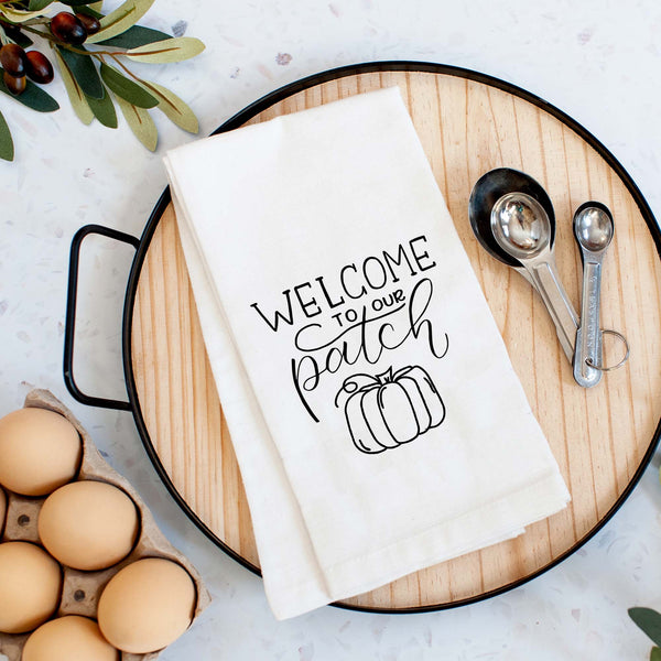 White floursack towel with black hand lettered illustrated design that says Welcome to our patch with pumpkin doodle shown folded on a serving tray with a set of measuring spoons and fresh eggs
