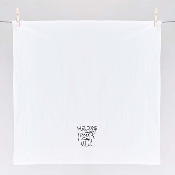 White floursack towel with black hand lettered illustrated design that says Welcome to our patch with pumpkin doodle shown unfolded and hanging from clothes pins