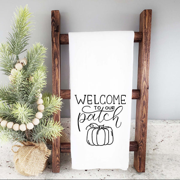 White floursack towel with black hand lettered illustrated design that says Welcome to our patch with pumpkin doodle shown folded and hanging from wooden display ladder and mini Christmas tree