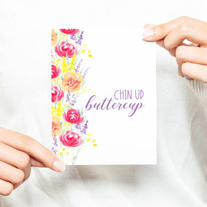 summery watercolor floral bouquets on an encouragement greeting card that says chin up buttercup with a white A2 envelope shown with a woman in a white sweater holding card