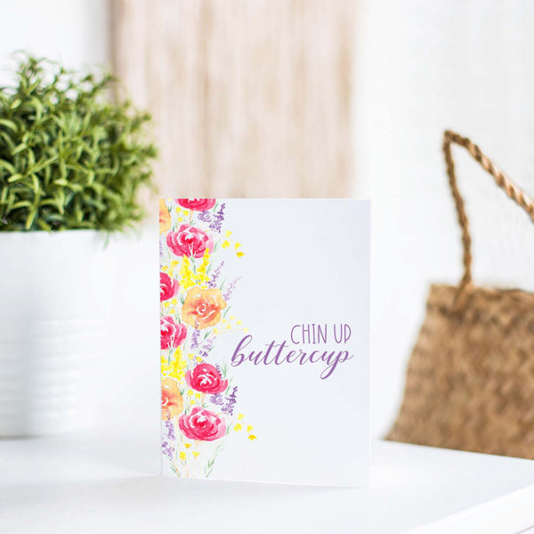 summery watercolor floral bouquets on an encouragement greeting card that says chin up buttercup with a white A2 envelope shown standing on a white table with a plant and handbag
