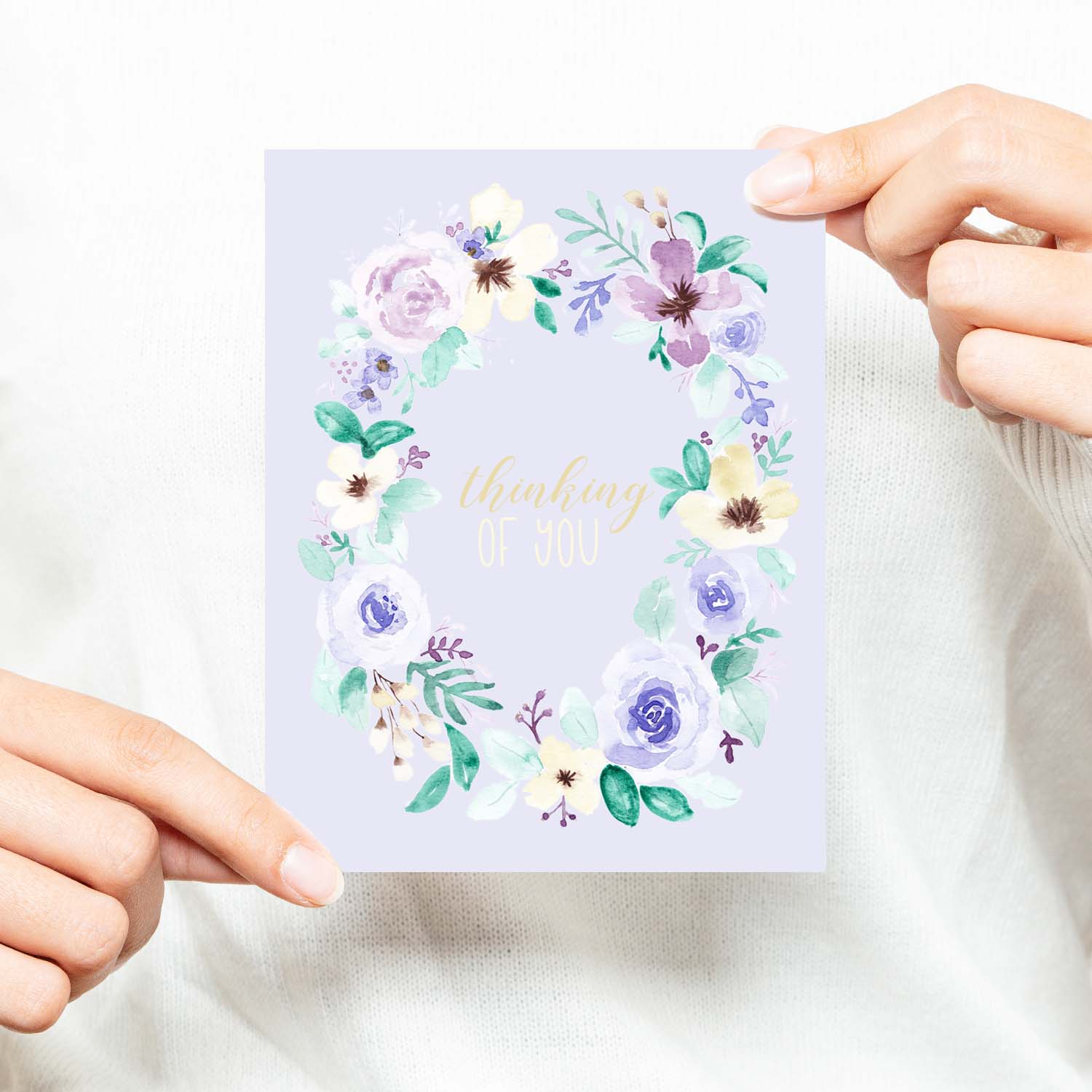watercolor wild flower wreath on a friendship greeting card that says thinking of you in the center with a white A2 envelope shown with a woman in a white sweater holding card